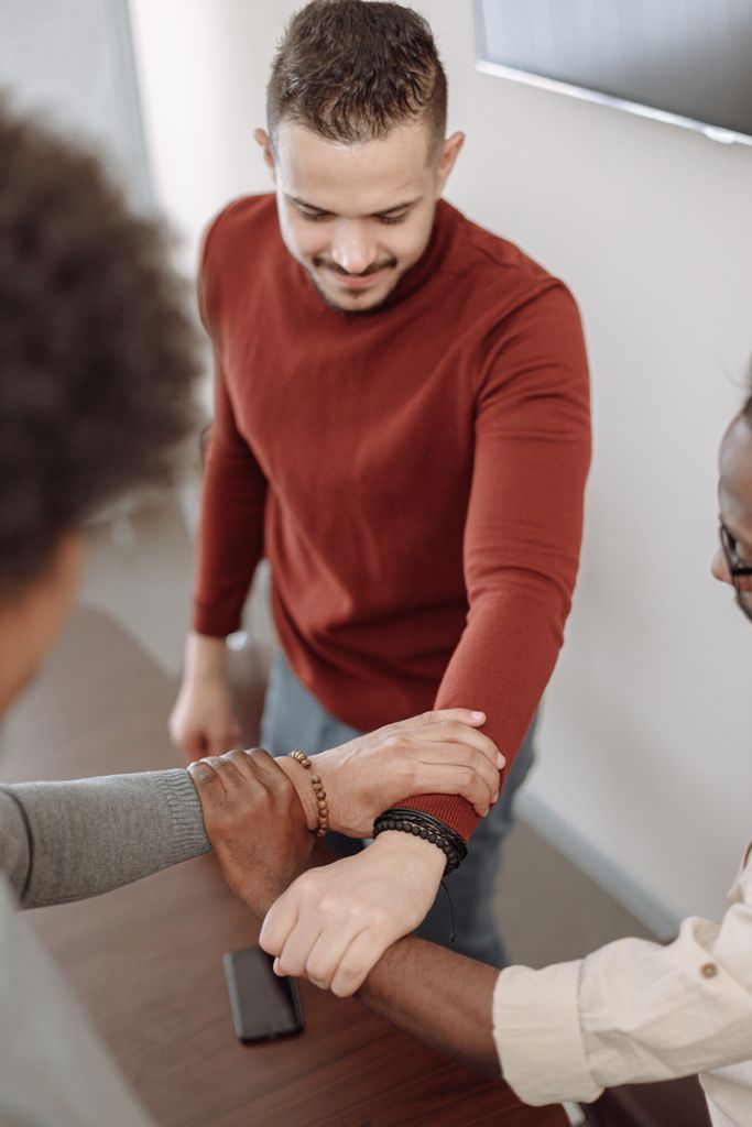 A diverse group of adults symbolizing teamwork by interlocking arms indoors.